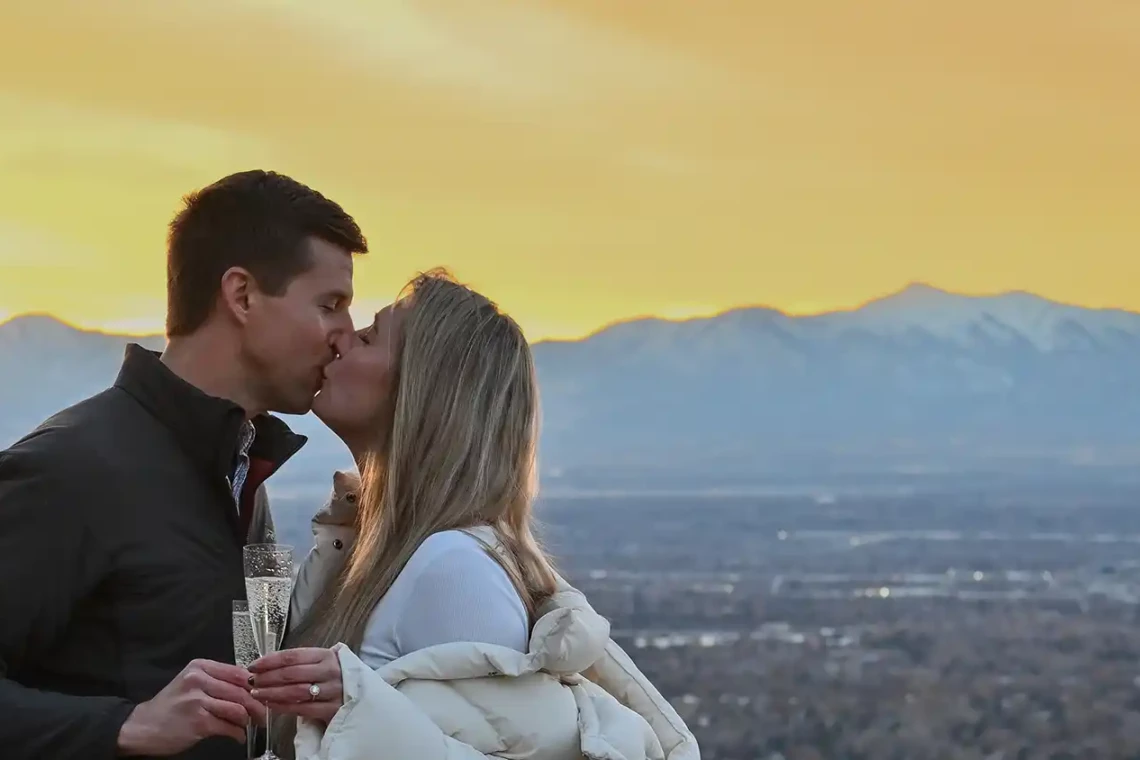 A newly engaged couple kissing at sunset with Salt Lake City as the background.
